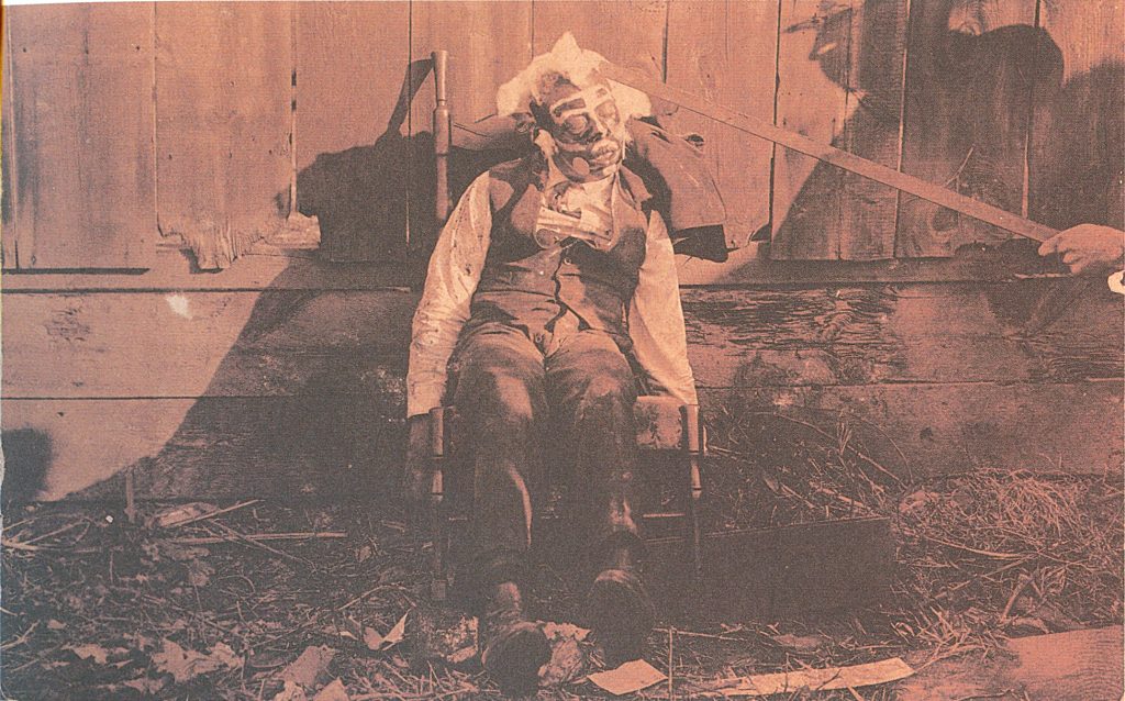 "The bludgeoned body of an African American male, propped in a rocking chair, blood-spattered clothes, white and dark paint applied to face, circular disks glued to cheeks, cotton glued to face and head, shadow of a man using rod to prop up the victim's head. Circa 1900, location unknown. Gelatin silver paint. Real photo postcard