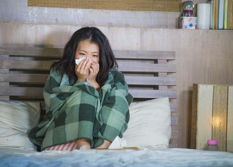 sweet Asian Korean woman in pajamas covered with blanket sick suffering cold and flu taking temperature with thermometer in bed feeling feverish blowing her nose coughing