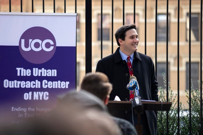 Council Member Ben Kallos smiling during his speach with "The Urban Outreach Center of NYC" banner behind him.