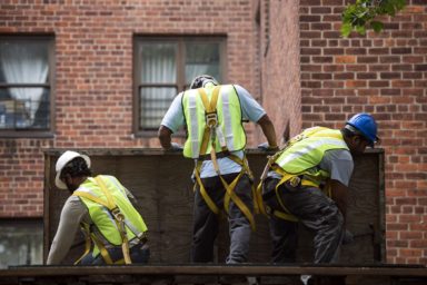 Three repairmen working on a plywood in front of a NYCHA building
