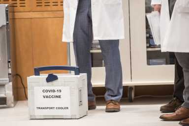 A medical container with a label that reads "COVID-19 vaccine Transport Cooler." on the floor