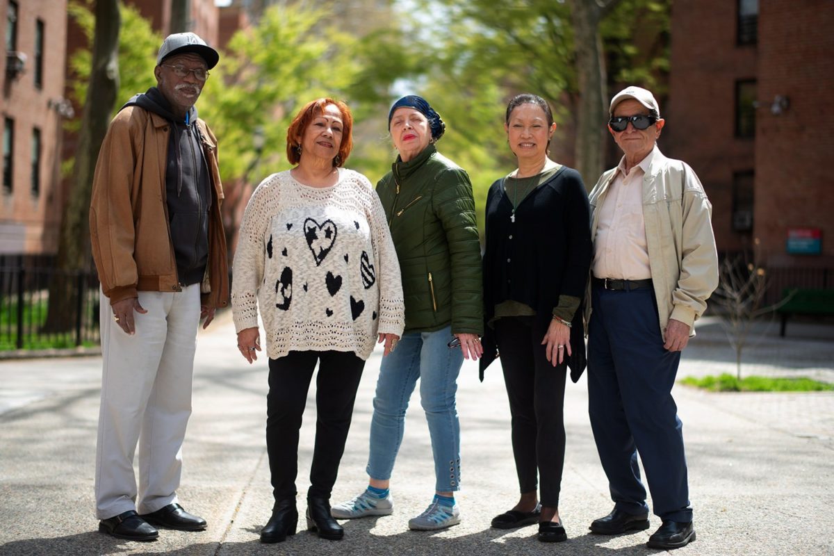 Five senior citizens, 2 males and 3 females, standing and looking at the camera surrounded by NYCHA building