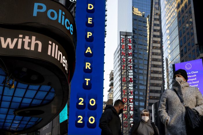 A view of Times Square with pedestrians and LED billboards, one that reads "Depart 2020" another one that reads "Police..(fragmented)" "with Illness..(fragmented)"