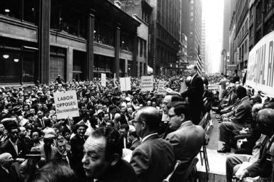 Charles Zimmerman speaks at a civil rights-labor movement rally in the New York Garment District on 38th Street near 7th Avenue in New York City. Signs include "Labor Opposes Discrimination"