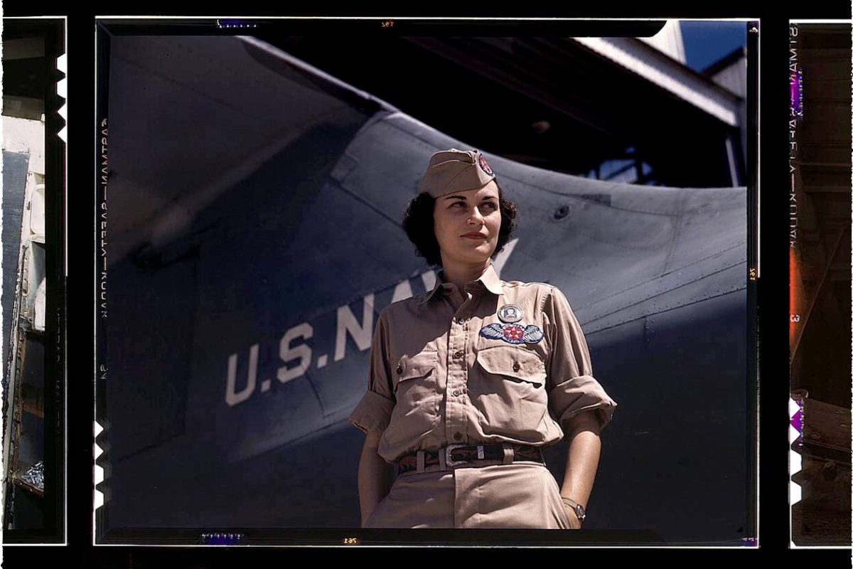 A young woman in U.S. Navy uniform looking sideway with her hands in pocket in front of a jet