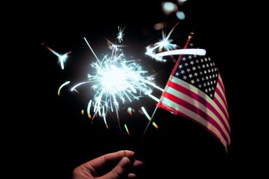 A hand holding a small american flag and a handheld firework in the dark